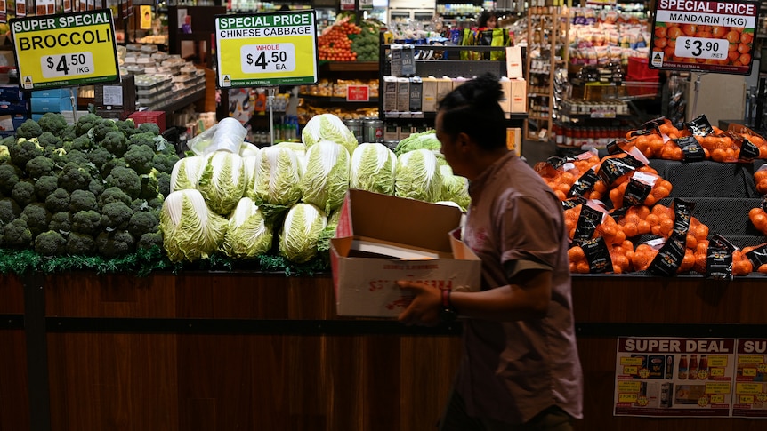 A person carrying a box walks past piled displays of vegetables