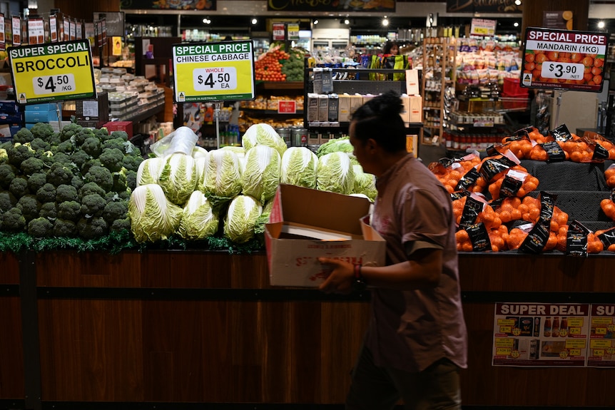 A person carrying a box walks past piled displays of vegetables