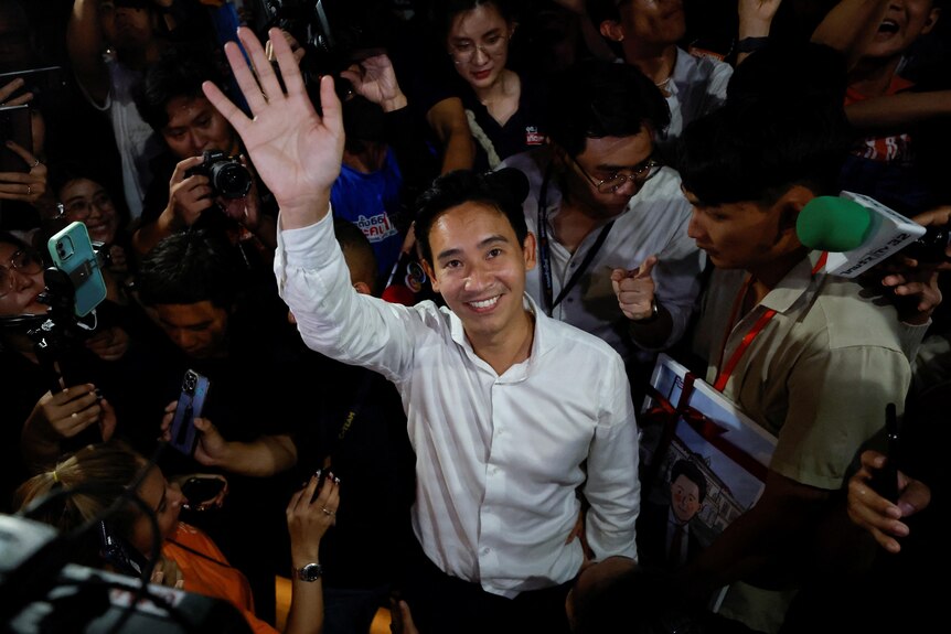 A young Thai man in white shirt smiles and waves to camera with people taking photos around him.