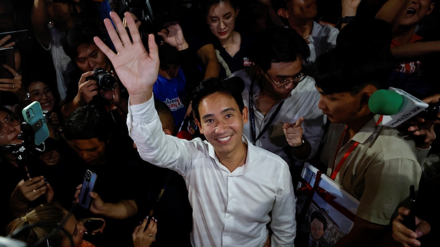 A young Thai man in white shirt smiles and waves to camera with people taking photos around him.