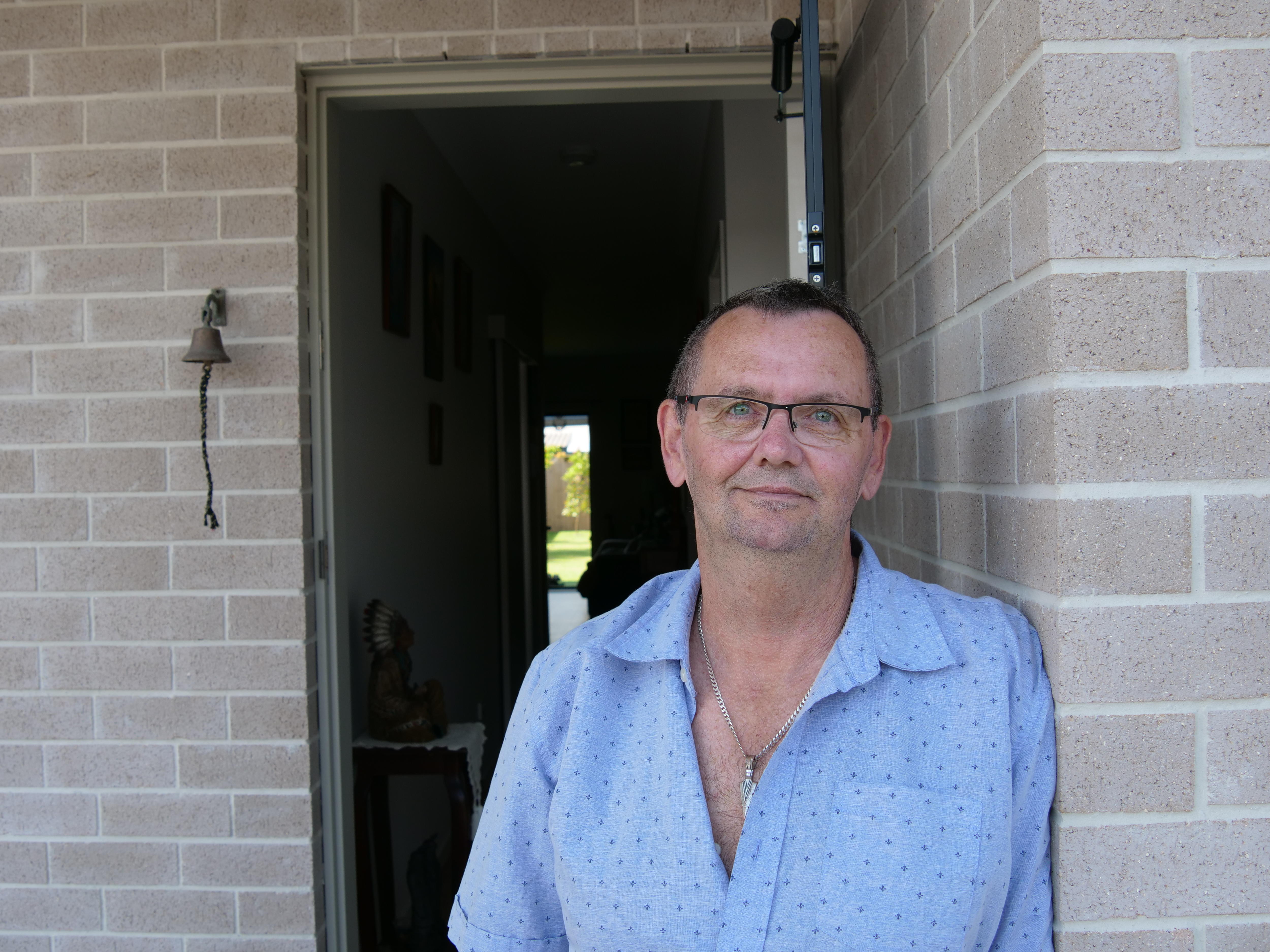 A middle-aged, bespectacled man leans against a brick wall next to the entrance of a flat.