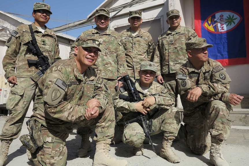 Seven soldiers of Micronesian appearance pose for a photo in US defence uniforms with a Guam flag hanging in the background.