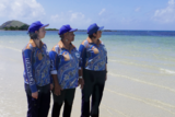 Three women wearing blue collared shirts and caps stand by the sea on a sunny day, looking out at clear waters