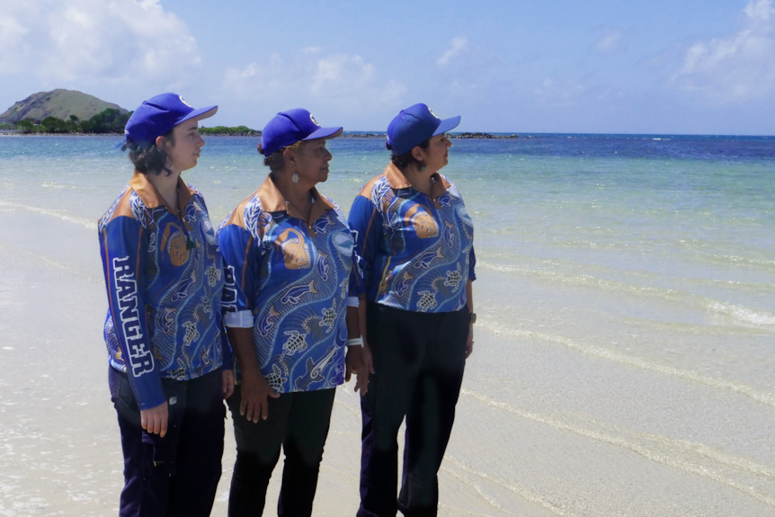 阳光明媚的日子里，三名身穿蓝色衬衫和帽子的女人站在海边，看着清澈的海水