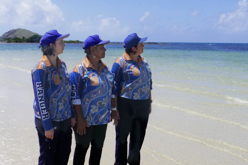 Three women wearing blue collared shirts and caps stand by the sea on a sunny day, looking out at clear waters