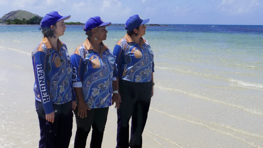 Three women wearing blue collared shirts and caps stand by the sea on a sunny day, looking out at clear waters