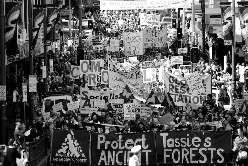 A black and white image showing a protest march filling a city street, with people holding banners urging protection of forests