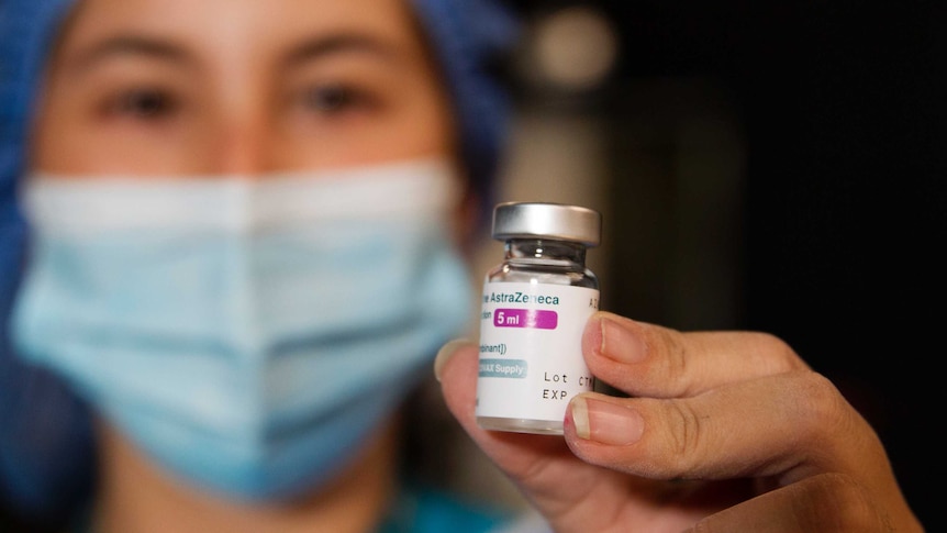 A nurse wearing blue scrubs and a light green mask holds up a vial of the AstraZeneca vaccine