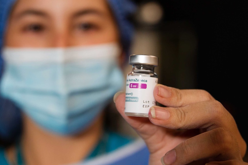 A nurse wearing blue scrubs and a light green mask holds up a vial of the AstraZeneca vaccine