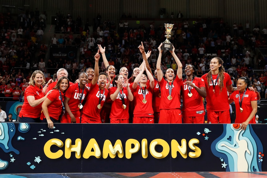 The USA women's basketball team stands behind a sign that says 'champions', they're cheering and one player raises the trophy.