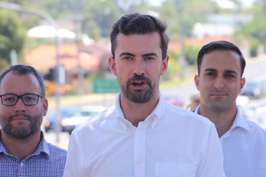 A close up of Zak Kirkup, centred, wearing a white shirt flanked by two other men, standing outside at a media conference.