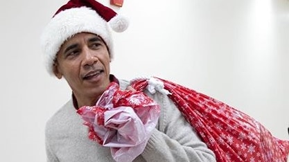 Barrack Obama wearing a Santa hat and carrying a red sack full of presents.