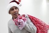 Barrack Obama wearing a Santa hat and carrying a red sack full of presents.
