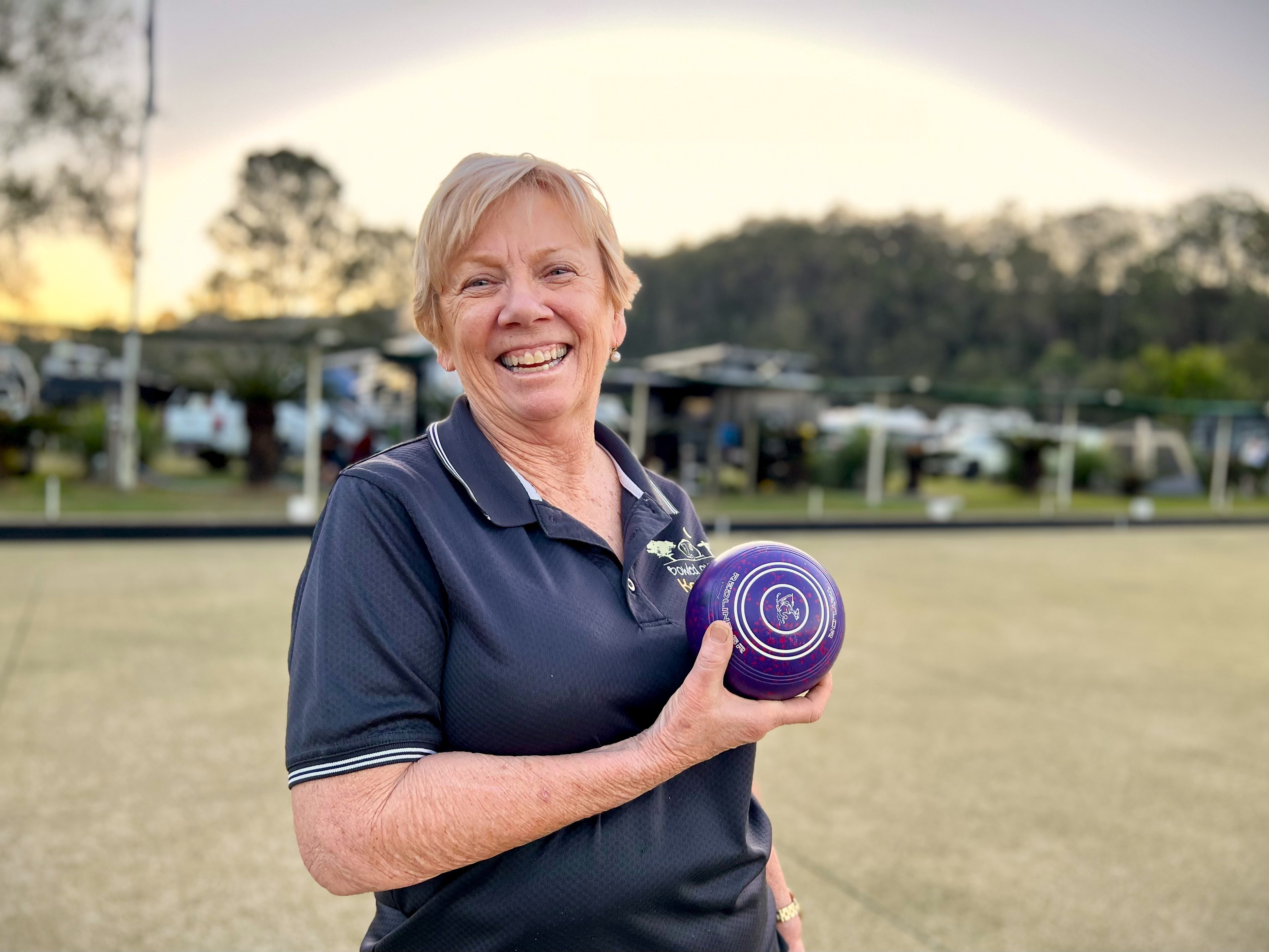 A smiling woman holds up a bowling bowl on the green, with caravans parked behind her.