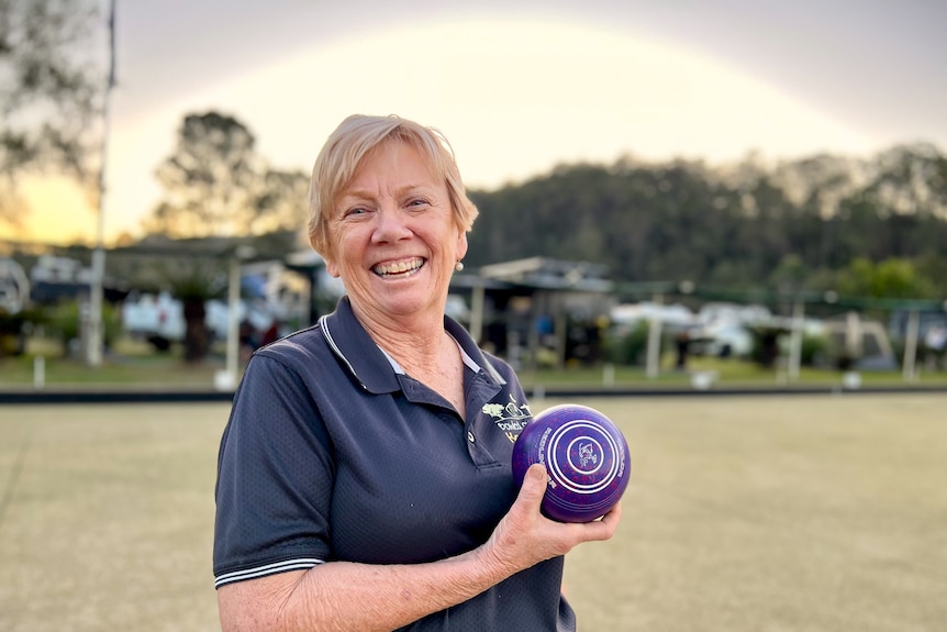 A smiling woman holds up a bowling bowl on the green, with caravans parked behind her.