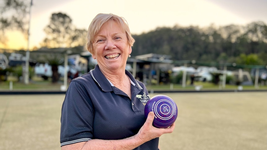 A smiling woman holds up a bowling bowl on a green. Caravans are parked nearby.
