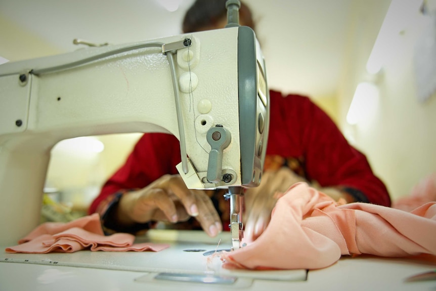 A woman's hands are seen as she holds a piece of pale pink fabric under a sewing machine. She wears red.