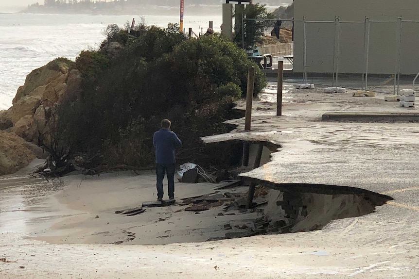 A badly eroded stretch of beach with a man standing on the sand in front of a car park that is overhanging it.