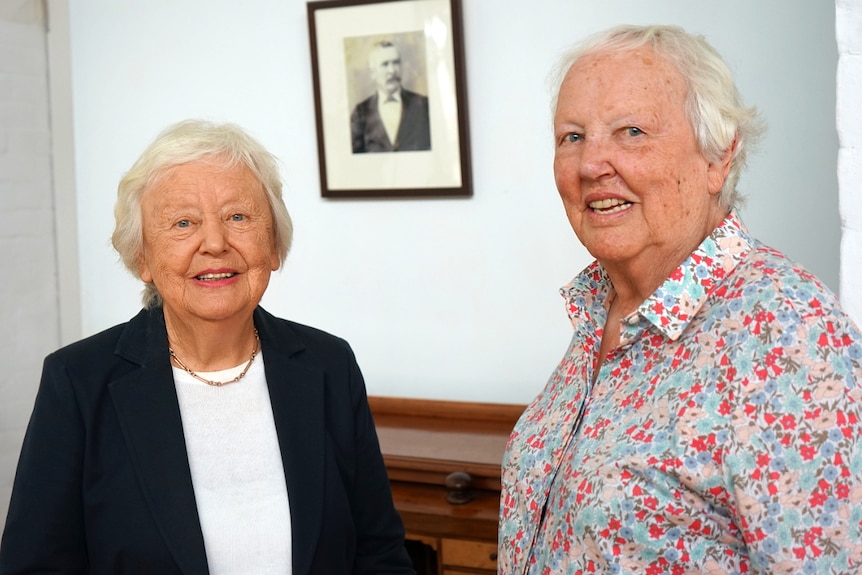 Two elderly ladies look at a camera with a portrait of their grandfather behind them