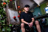 A slightly smiling Asian man in black t-shirt, shorts, cap sitting in a restaurant with red Chinese lanterns, plants.