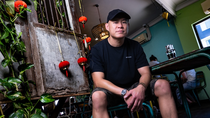 A slightly smiling Asian man in black t-shirt, shorts, cap sitting in a restaurant with red Chinese lanterns, plants.