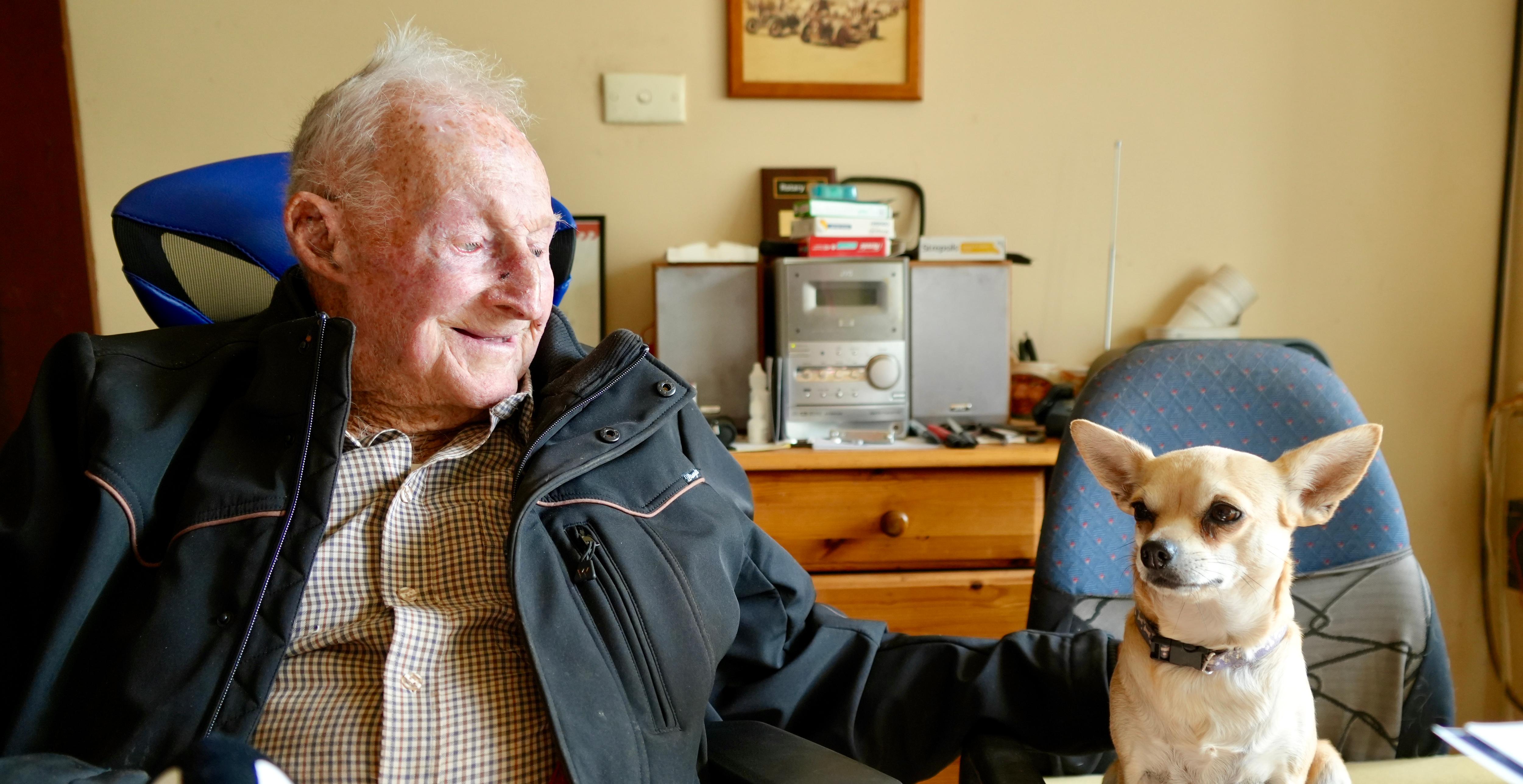 a man sitting in a chair with a black jacket patting a small yellow dog sitting next to him 