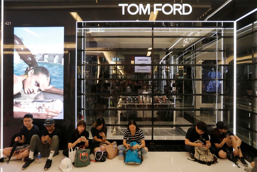 A group of people sit on the floor outside a closed Tom Ford boutique