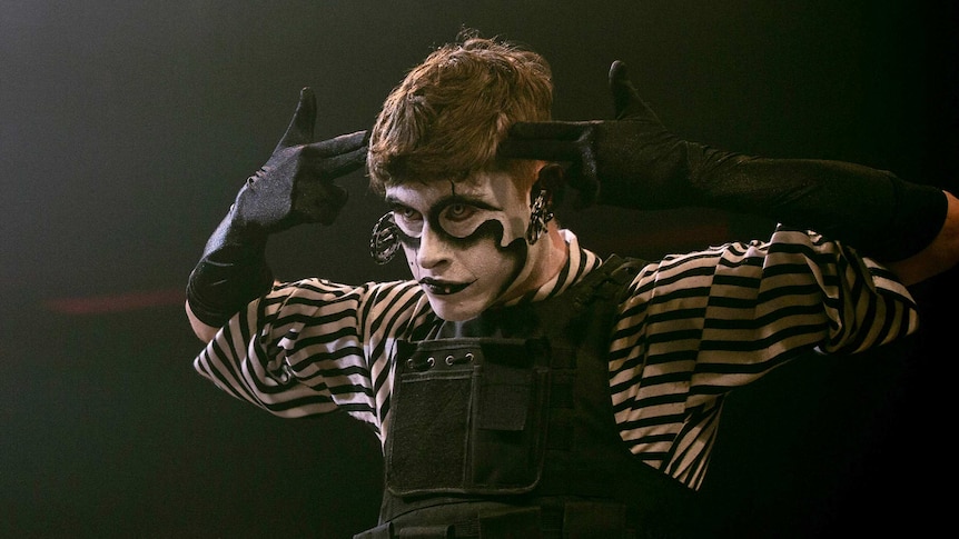 A man in black-and-white face paint and black gloves gestures to his head on stage.