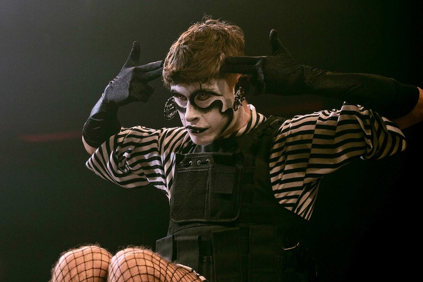 A man in black-and-white face paint and black gloves gestures to his head on stage.