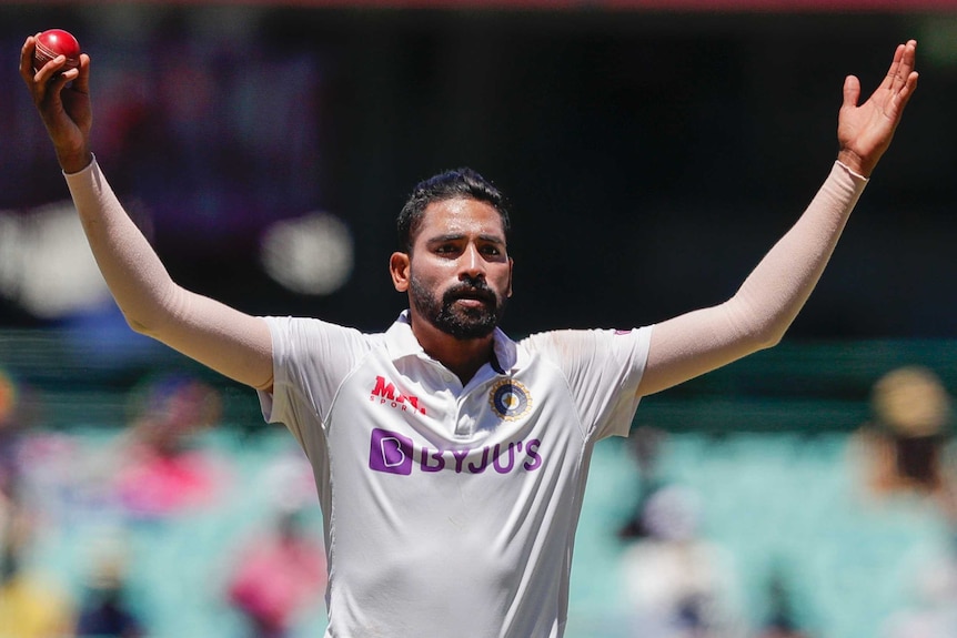 Mohammed Siraj holds his hands out wide above his head, holding a red cricket ball in his right hand