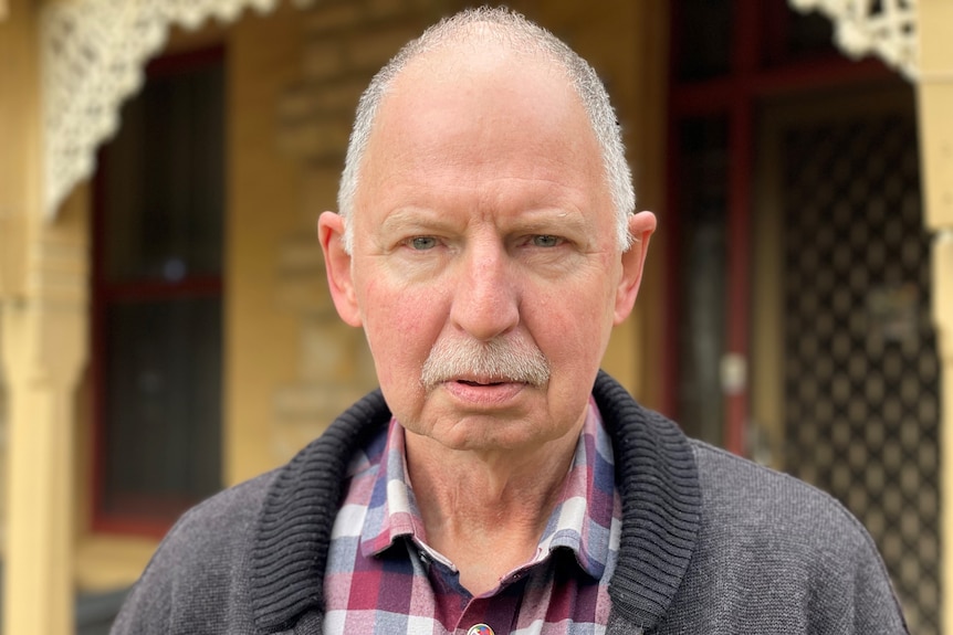 An elderly man wearing a check shirt standing in front of a house