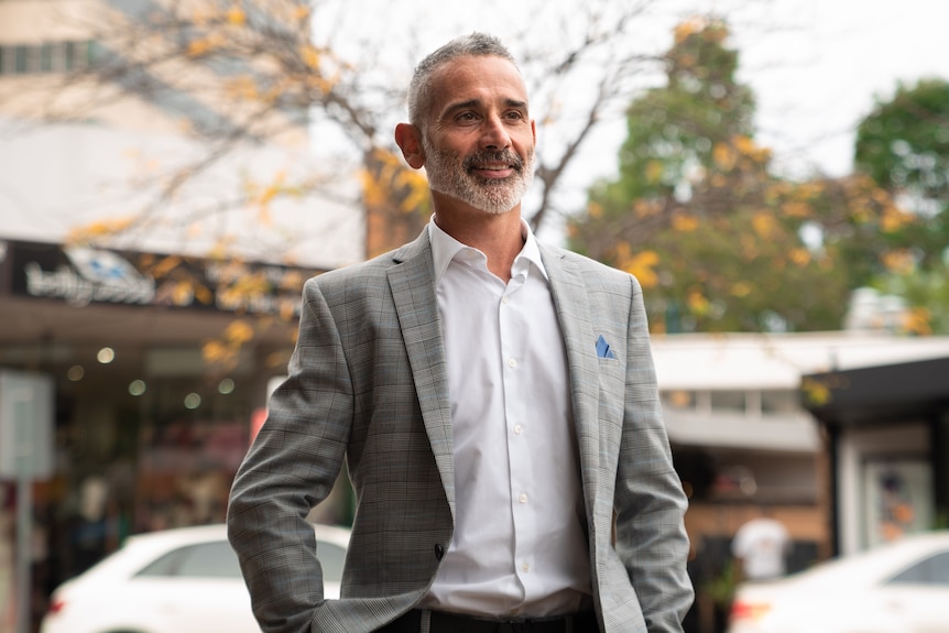 A man wearing a grey suit and white shirt stands on a town street. 