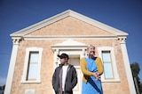 A man and a woman stand in front of an old hall looking in opposite directions.