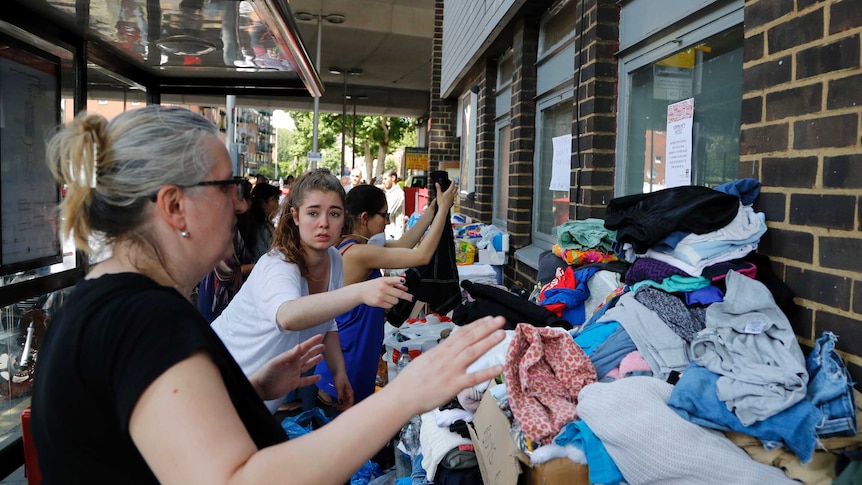 Women sort through piles of clothing stacked on tables outside a building