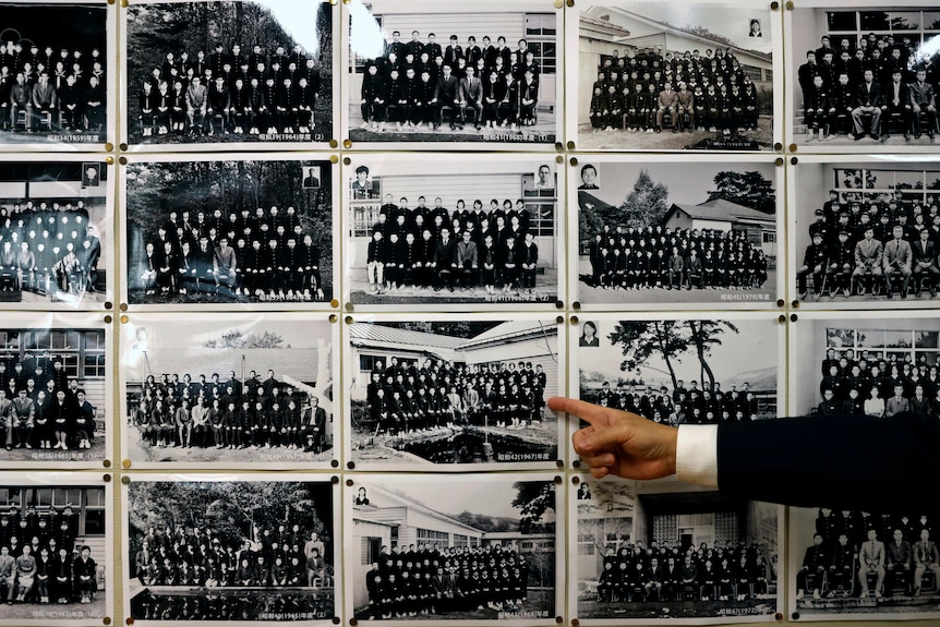 Yumoto Principal Mikio Watanabe points at a wall of black and white graduation photos from the school