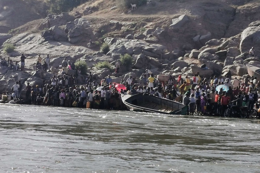 Hundreds of people stand on a river bank next to a small boat