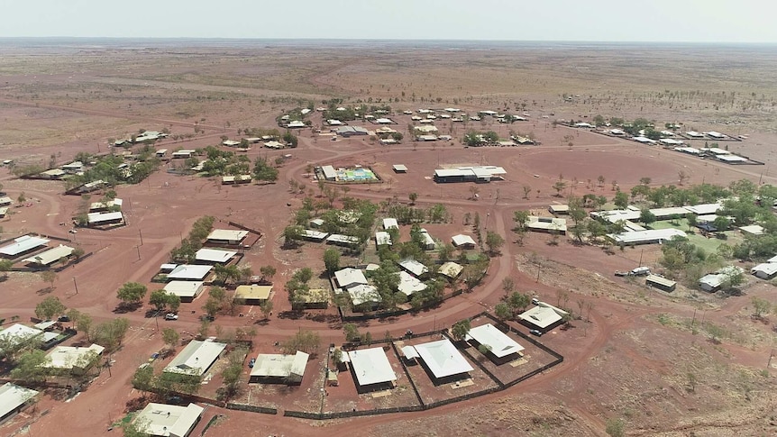 An aerial picture of a small town in the outback with red hills and sparse green shrubs.