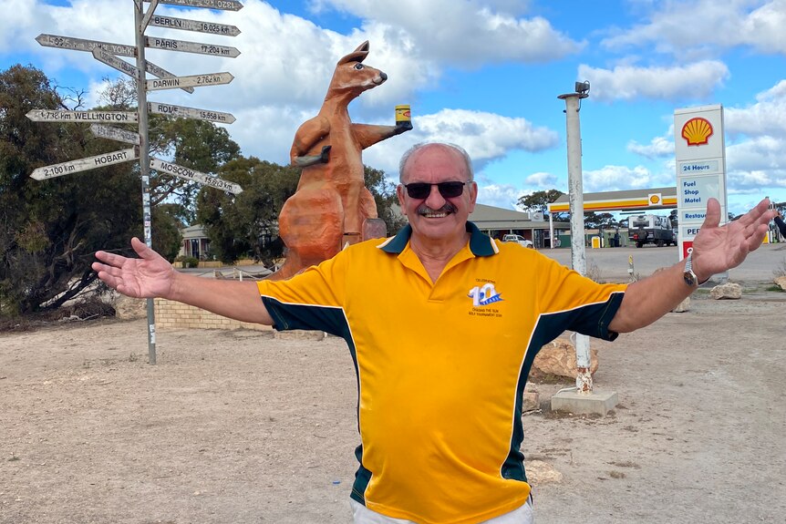 Man in yellow shirt with both arms outstretched in from of large Kangaroo, sign post with lots of direction signs and roadhouse 