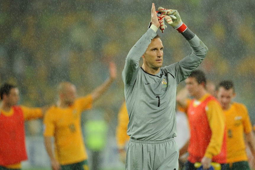Mark Schwarzer lifts his arms in the air wearing a grey goalkeeper's outfit with several Australian soccer players behind.
