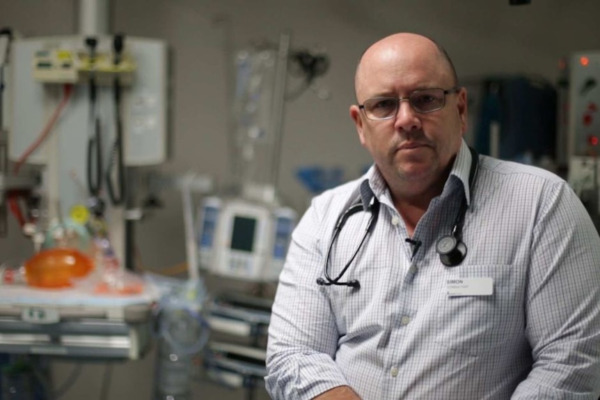 Man in shirt with stethoscope wrapped around neck looks at camera. He's in a hospital setting.