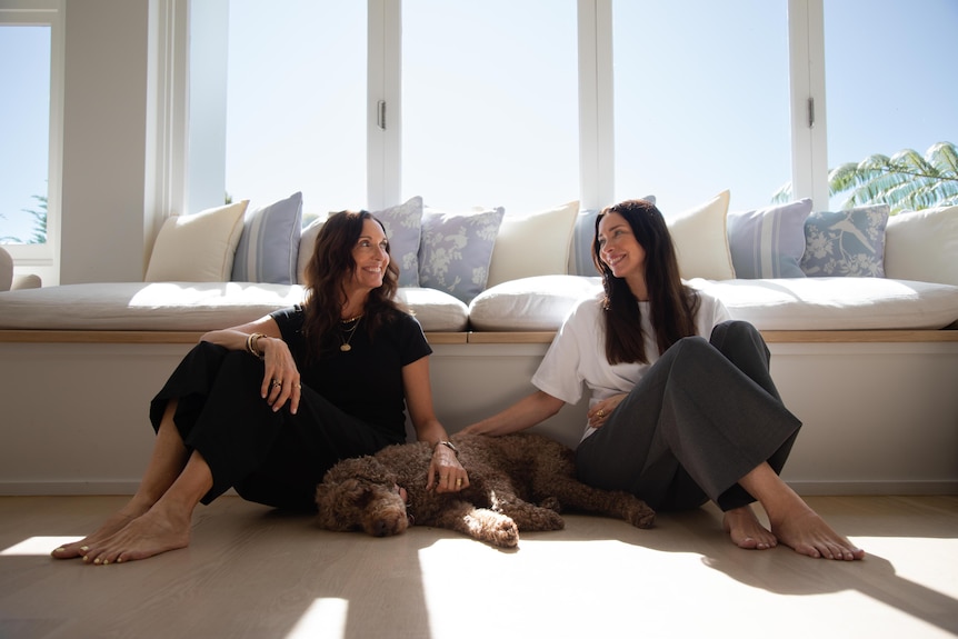 Two brunette women sit on the floor of a lounge room looking at each other smiling while patting a dog that lay on the ground