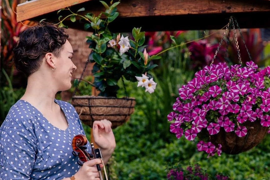 Sophia mackson in a blue dress holding a viola looking away from the camera with hanging flower pot and bushes in background