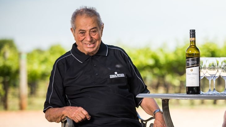 An old man in a navy polo top and slacks sits at an outdoor table with a wine bottle on it in a vineyard on a sunny day.
