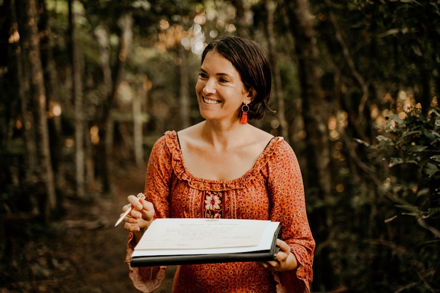 A smiling woman wearing an orange dress stands in a bush clearing holding a register and a pen.