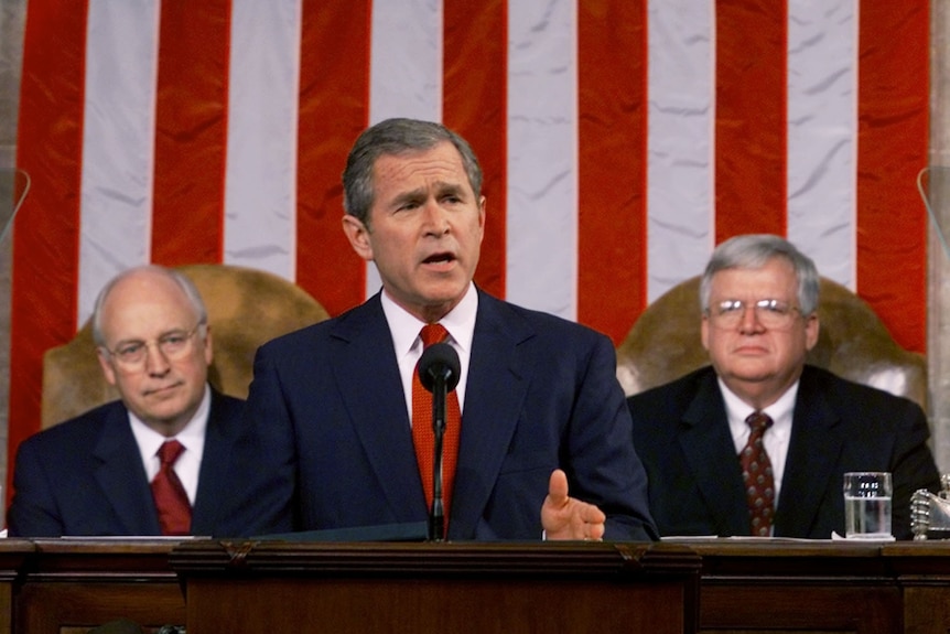 George W Bush speaks in front of the American flag while Dick Cheney and Dennis Hastert look on.