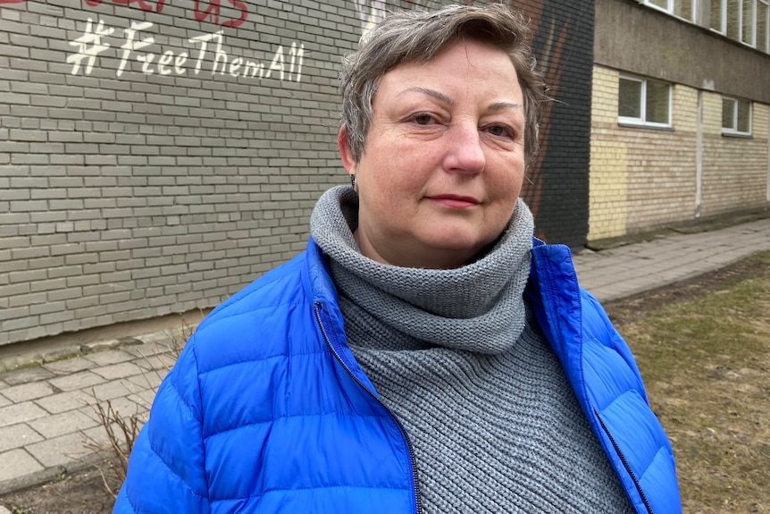 A woman in a grey turtleneck and blue puffer looks at the camera. graffiti on a wall behind her reads 'free them all'