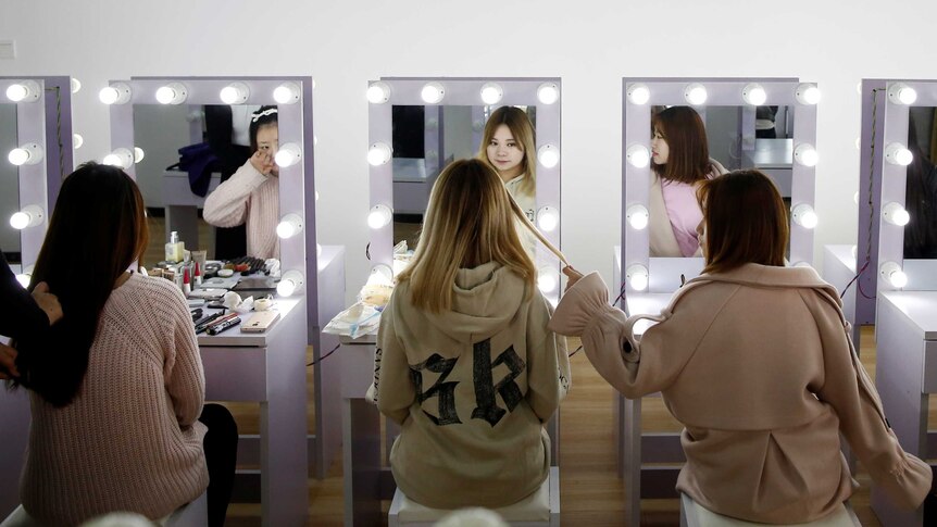 Three girls sit in front of make-up mirrors.
