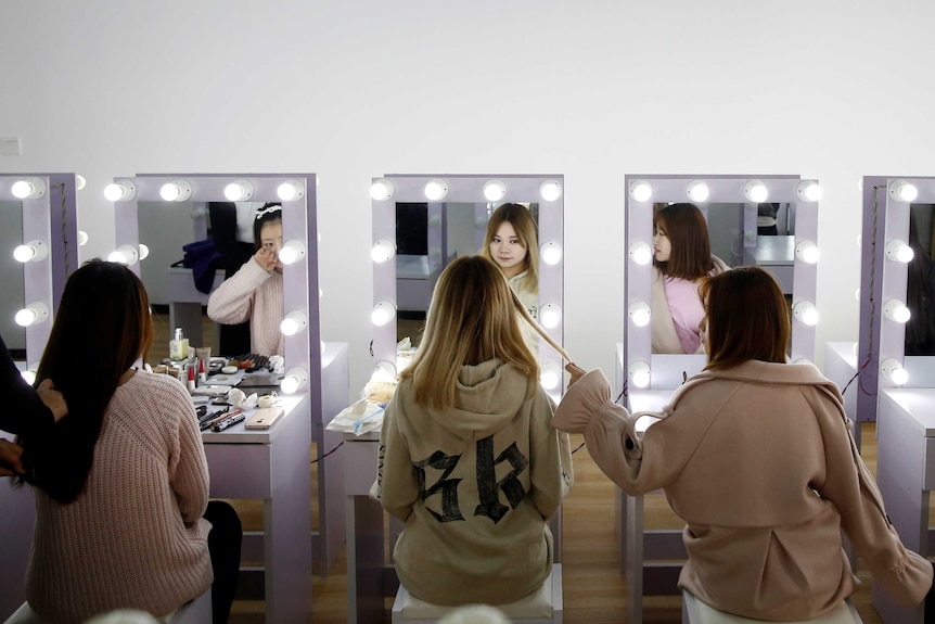 Three girls sit in front of make-up mirrors.