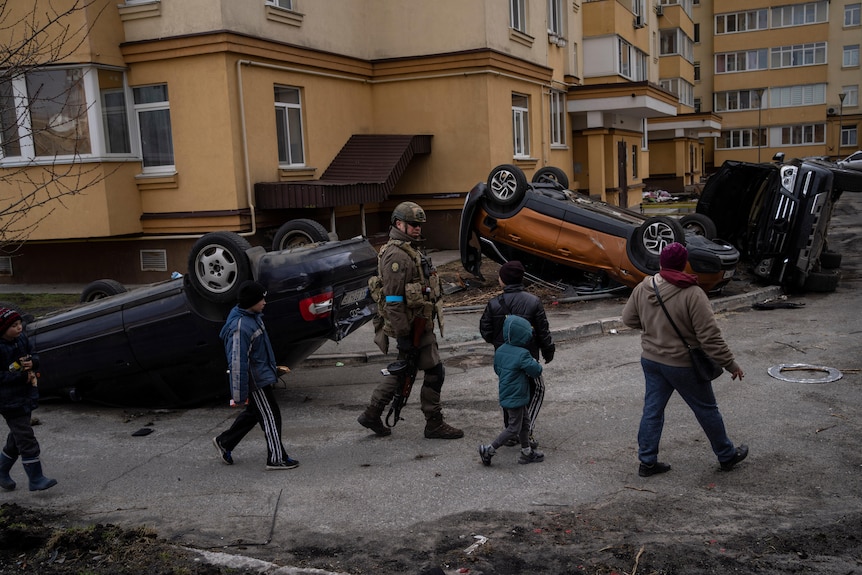 A soldir walking along with children passing destroyed cars.