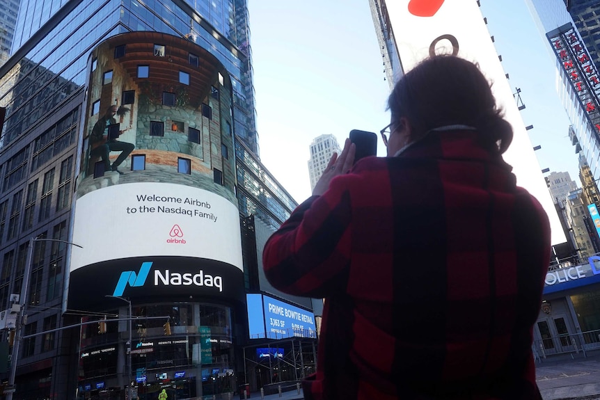 A person takes a photo on their phone of the Nasdaq digital sign showing Airbnb logo.